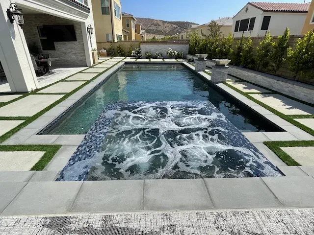 A modern rectangular swimming pool with a built-in hot tub area in the foreground. The pool is surrounded by a stone walkway, artificial grass, and residential homes in the background.